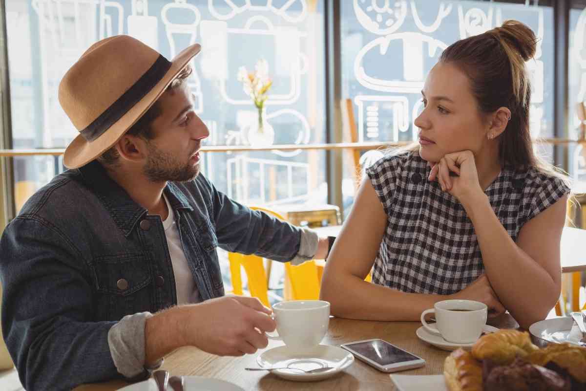 A couple sitting at a cafe table mid-conversation, one listening with quiet concern, evoking the difficult talk the Eight of Swords keeps postponing